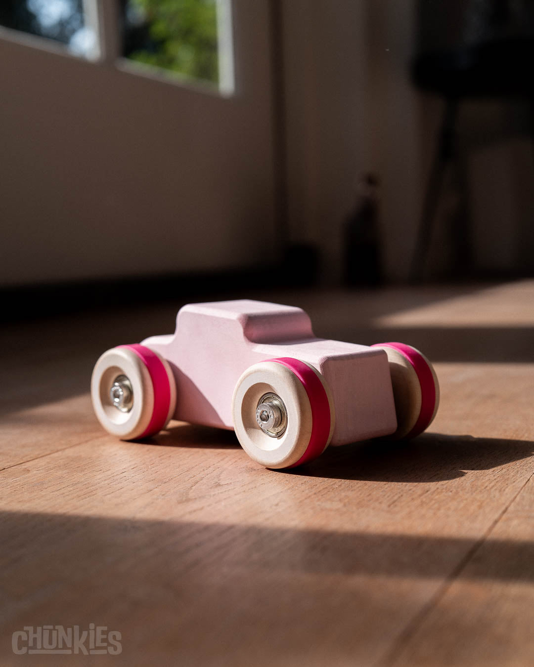Chill Pepper Chunkies No04  Wooden Toy Car The Hot Rod Perfectly Pink in a living room on a wooden floor and a blurred background. The sunlights shines through the the windows accentuating the colors and curvy shapes