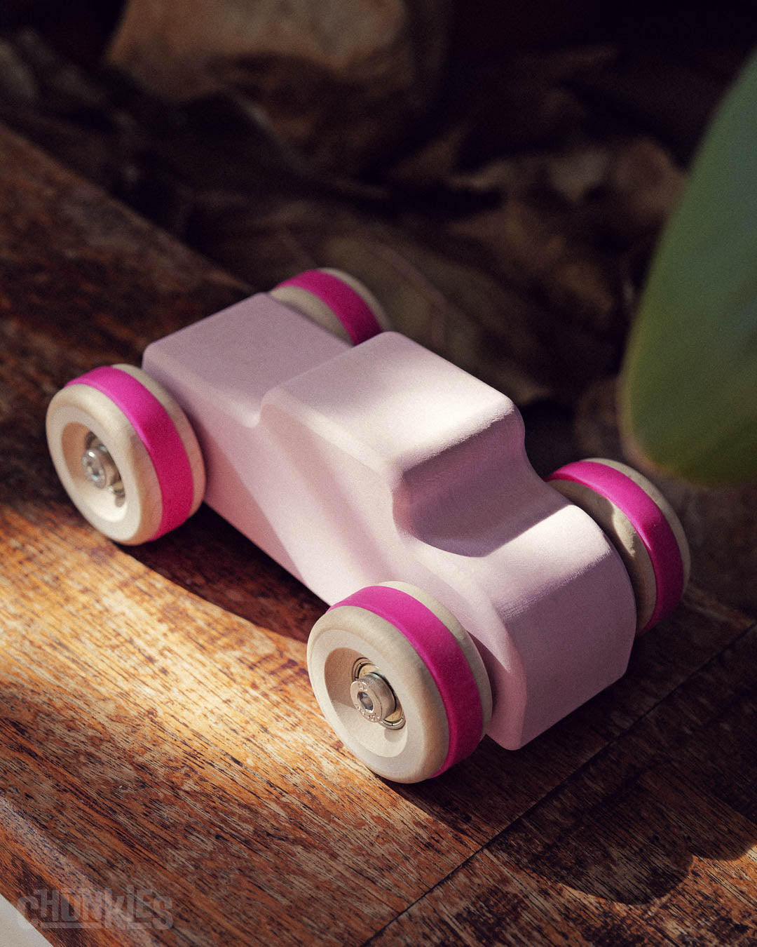 Top view of a Chill Pepper Chunkies No04  Wooden Toy Car The Hot Rod Perfectly Pink on a rustic wooden board and plants in the background. Made from sustainable maple wood in Germany and hand finished and assembled in The Netherlands