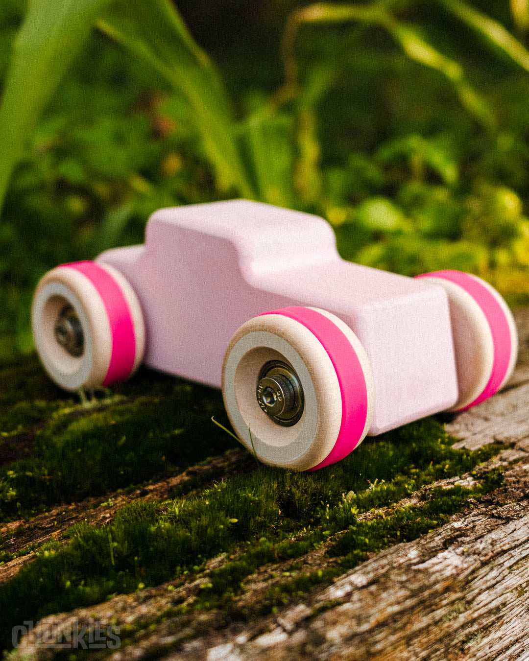 Outdoor shot of the Chill Pepper Chunkies No04  Wooden Toy Car The Hot Rod Perfectly Pink. It's sat on a rustic beam with moss and plants in the back