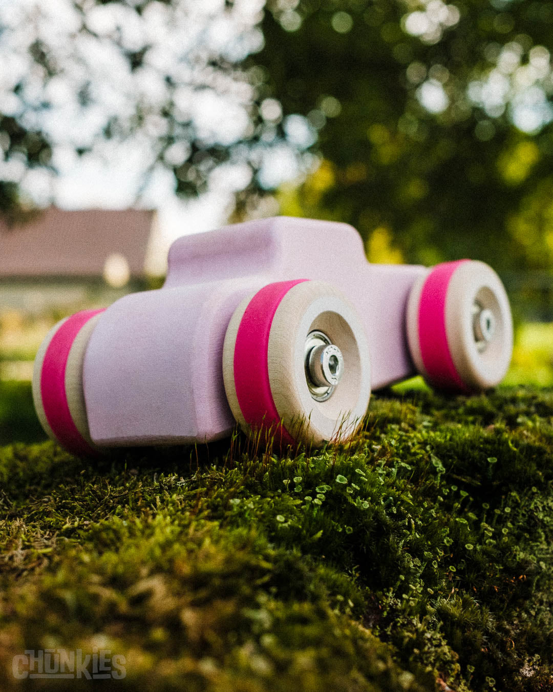 Outdoor back 3/4 shot of the Chill Pepper Chunkies No04  Wooden Toy Car The Hot Rod Perfectly Pink sitting on moss with trees in the back