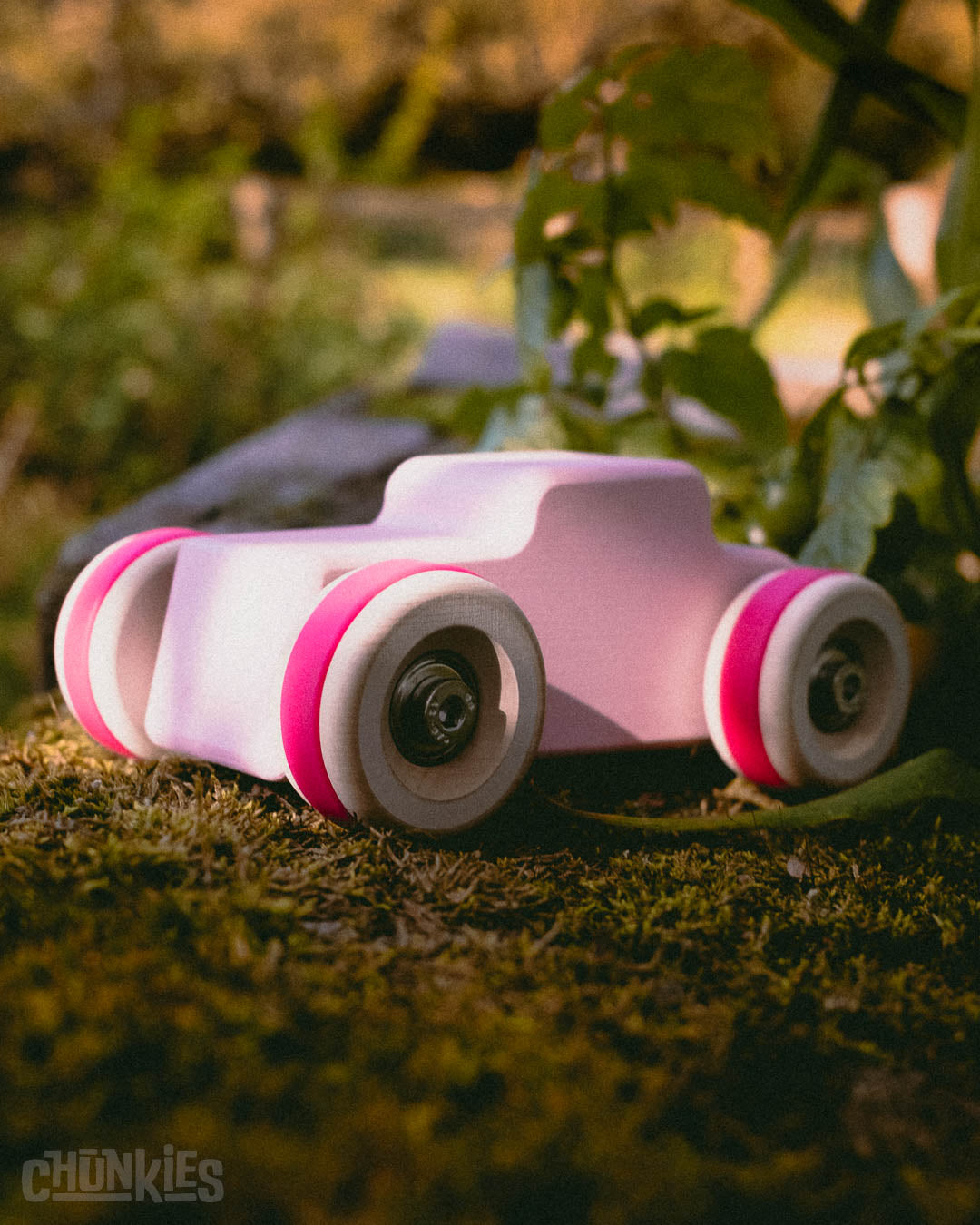 Outdoor shot of a Chunkies pink hot rod toy car made by Chill Pepper. The pink tires are on dark green moss with plants in the back to accentuate the natural materials of this child friendly and sustainable wooden toy car.