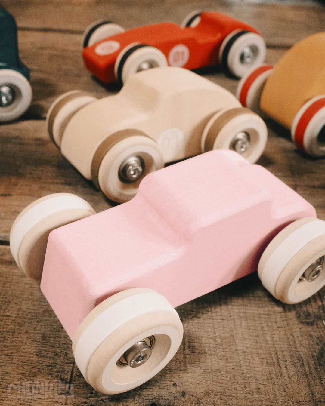 Top 3/4 view of the Chill Pepper Chunkies No04  Wooden Toy Car The Hot Rod Perfectly Pink with white tires and other Chunkies cars in the background. 