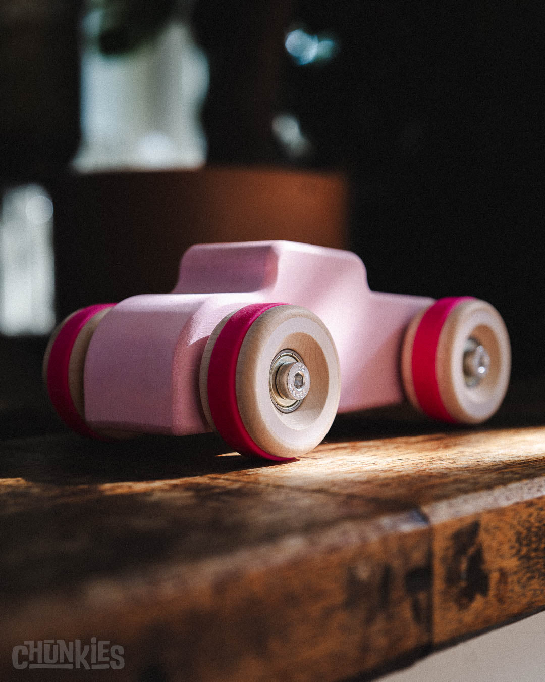 3/4 back view of the curvy back side of the Chill Pepper Chunkies No04  Wooden Toy Car The Hot Rod Perfectly Pink. Here on a rustic wooden ledge with plants in the background. The light pink water based and child friendly finish is matte and feels great in the hand