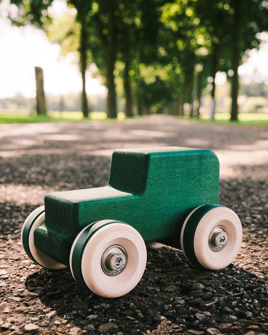 Green toy car with big wooden wheels on a gravel path with trees in the background. Solid Maple form Europe. Made in Germany. Model: Chill Pepper Chunkies No05 Wooden Toy Car The Explorer All-Terrain