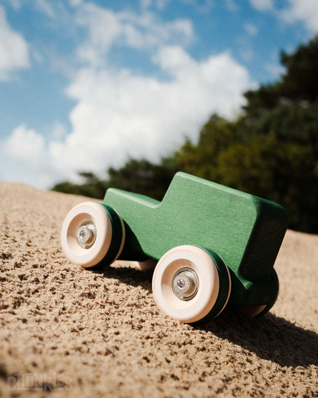 Green wooden toy car on a sandy surface with a blue sky and trees in the background made by Chill Pepper. Model: Chunkies No05 Wooden Toy Car The Explorer All-Terrain