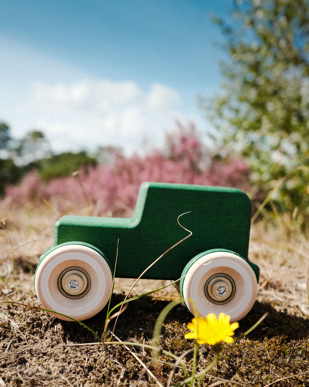 Wooden toy all-terrain car (The Explorer) on a grassy field with a yellow flower made by Chill Pepper in The Netherlands