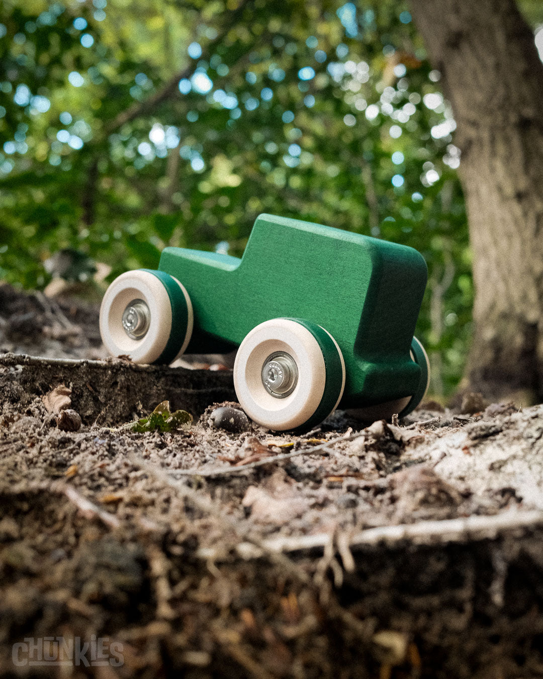 Green toy car with big wooden wheels on a forest floor. This is model Chill Pepper Chunkies No05 Wooden Toy Car The Explorer All-Terrain in Gorgeous Green matte chilfriendly stain