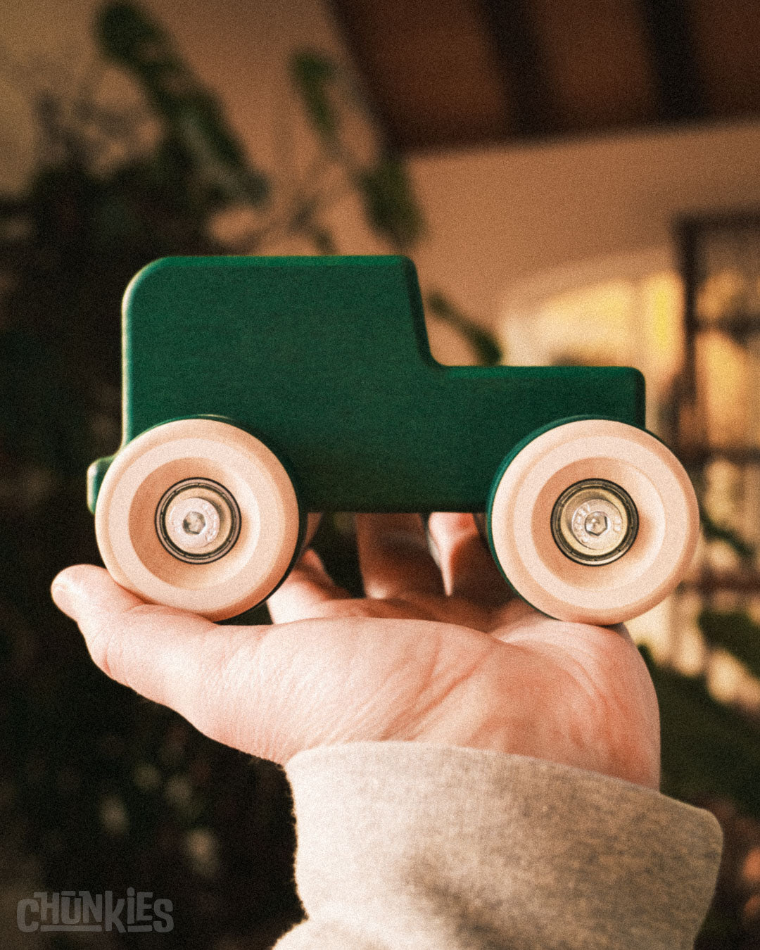 Chill Pepper Chunkies No05 Wooden Toy Car The Explorer All-Terrain - Hand holding a big green wooden toy truck with big wooden wheels against a blurred indoor background. A smooth ride is guaranteed by it's big wooden wheels, steel hardware and real skate bearings