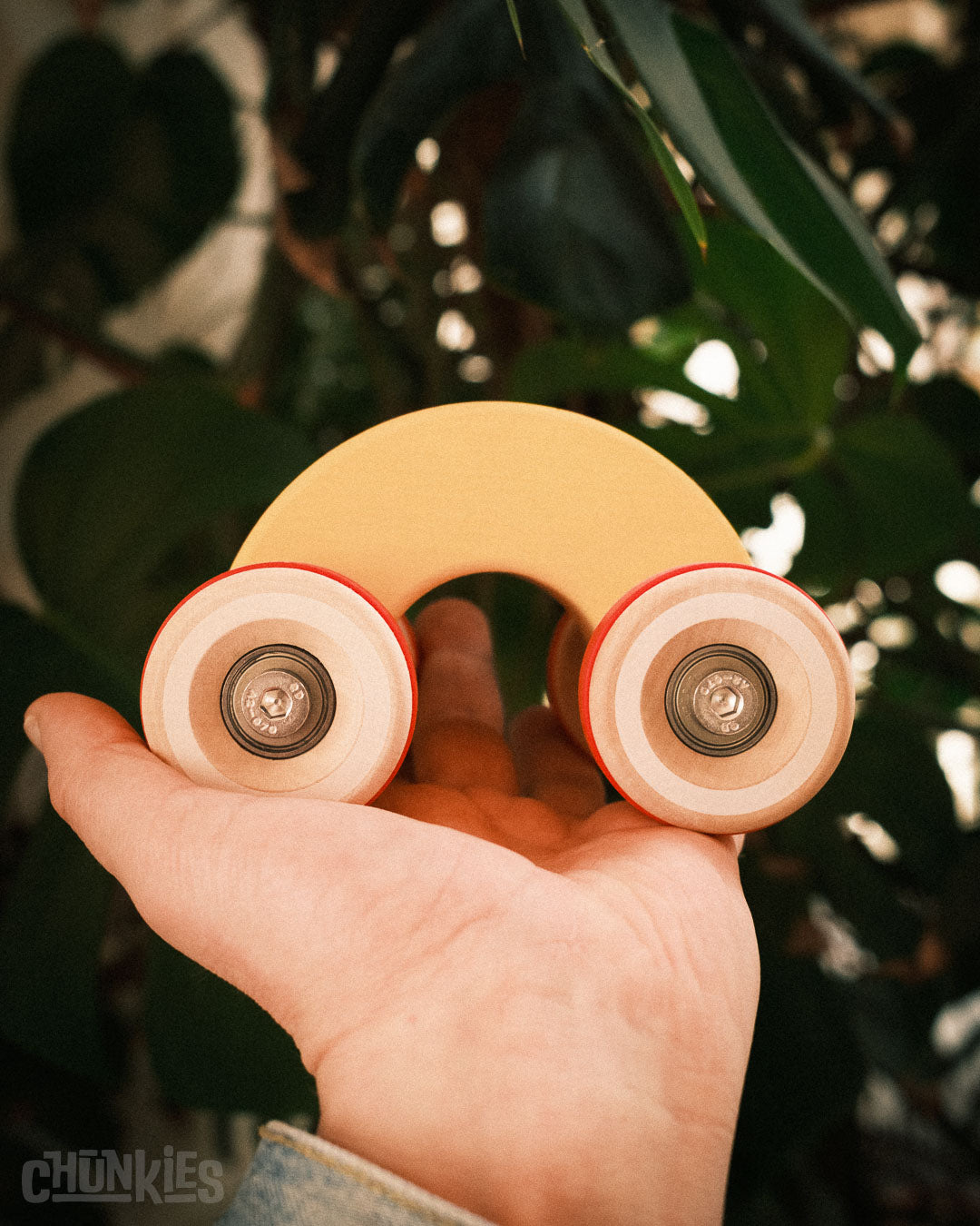 Hand holding a Chunkies wooden toy car with a blurred green leafy background. This is model: Chill Pepper Chunkies No06 Wooden Toy Car The Rainbow