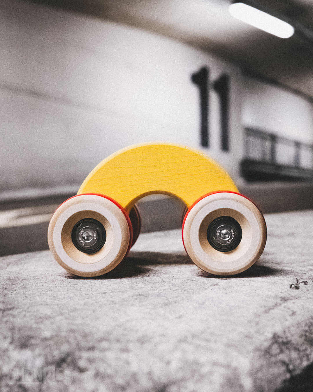 Side view of the Chill Pepper Chunkies No06 Wooden Toy Car The Rainbow in a parking garage. The yellow and red colors contrast nicely with the urban environment, but will look good in any interior too.