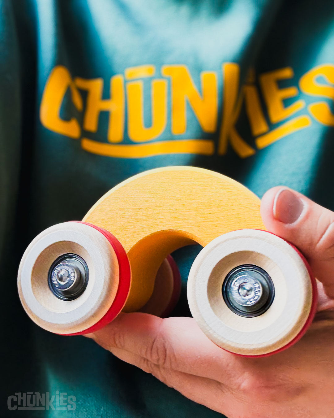 Hand held shot of the Chill Pepper Chunkies No06 Wooden Toy Car The Rainbow with a teal hoody in the background with the big bold yellow Chunkies logo printed on it