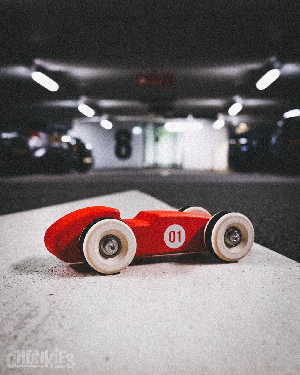 A big red wooden toy race car made of European maple wood with big wooden wheels and real skate bearings for a smooth ride. In the background a urban parking garage to set the scene. This big little car will be your child's favorite and will join you on every trip. Ready for big adventures and planet friendly