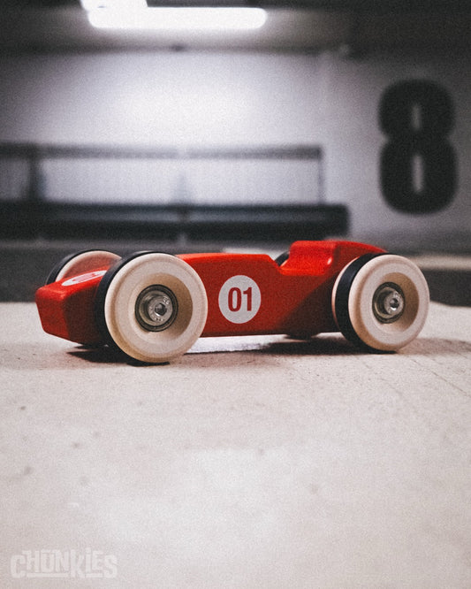 Chunkies No. 01 Wooden Racing Car "The 1956 Legend" in Radiant Red - 3/4 front view in urban garage setting. It's real skate bearings in made in Europe maple solid wooden body and wheels make this a pretty chunky toy car. You can feel the quality. Made for this and the next generations. A true heirloom. Not just for kids, but also for car enthousiasts to add to their collection