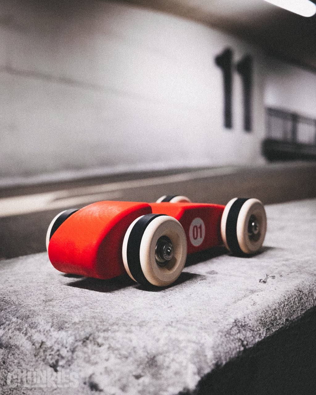 Rear view of red wooden toy race car showing robust European Maple construction. Big wheels and real skate bearings. Shown here in an urban parking garage. The child friendly water based paint has a matte texture. Together with the rounded edges it feels great to hold. This big little car will go on any adventure: on holiday, to school, to a friends house and to sleepovers and the grandparents.