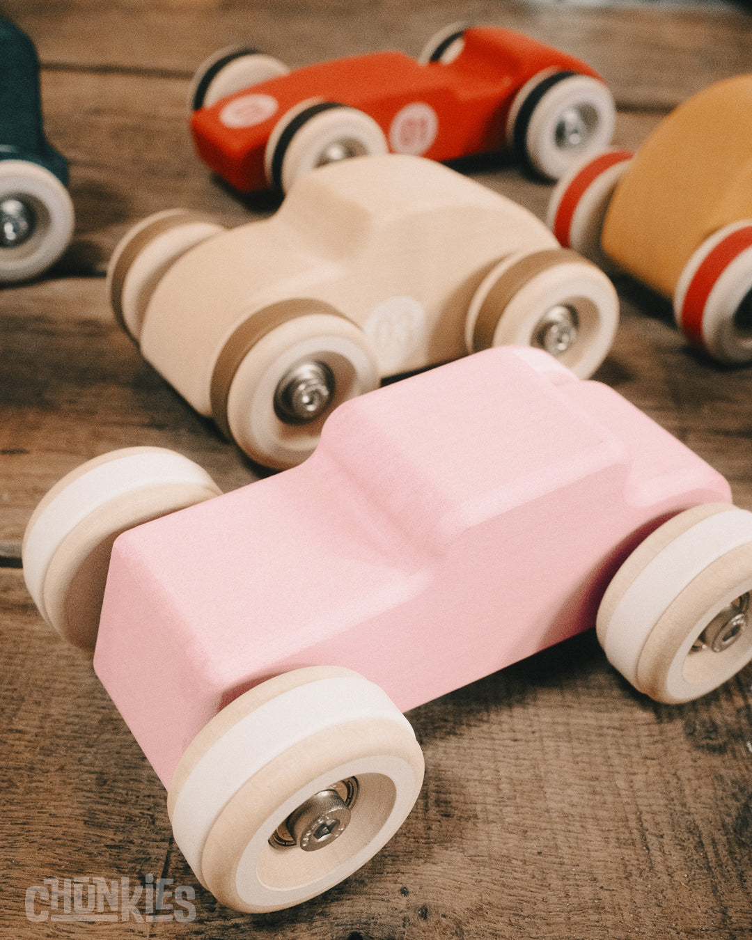 Various colorful Chunkies wooden toy cars shown on a wooden table. In the front  The pink hot rod. In the middle the natural wooden Bug and in the back the Chunkies Legend racer