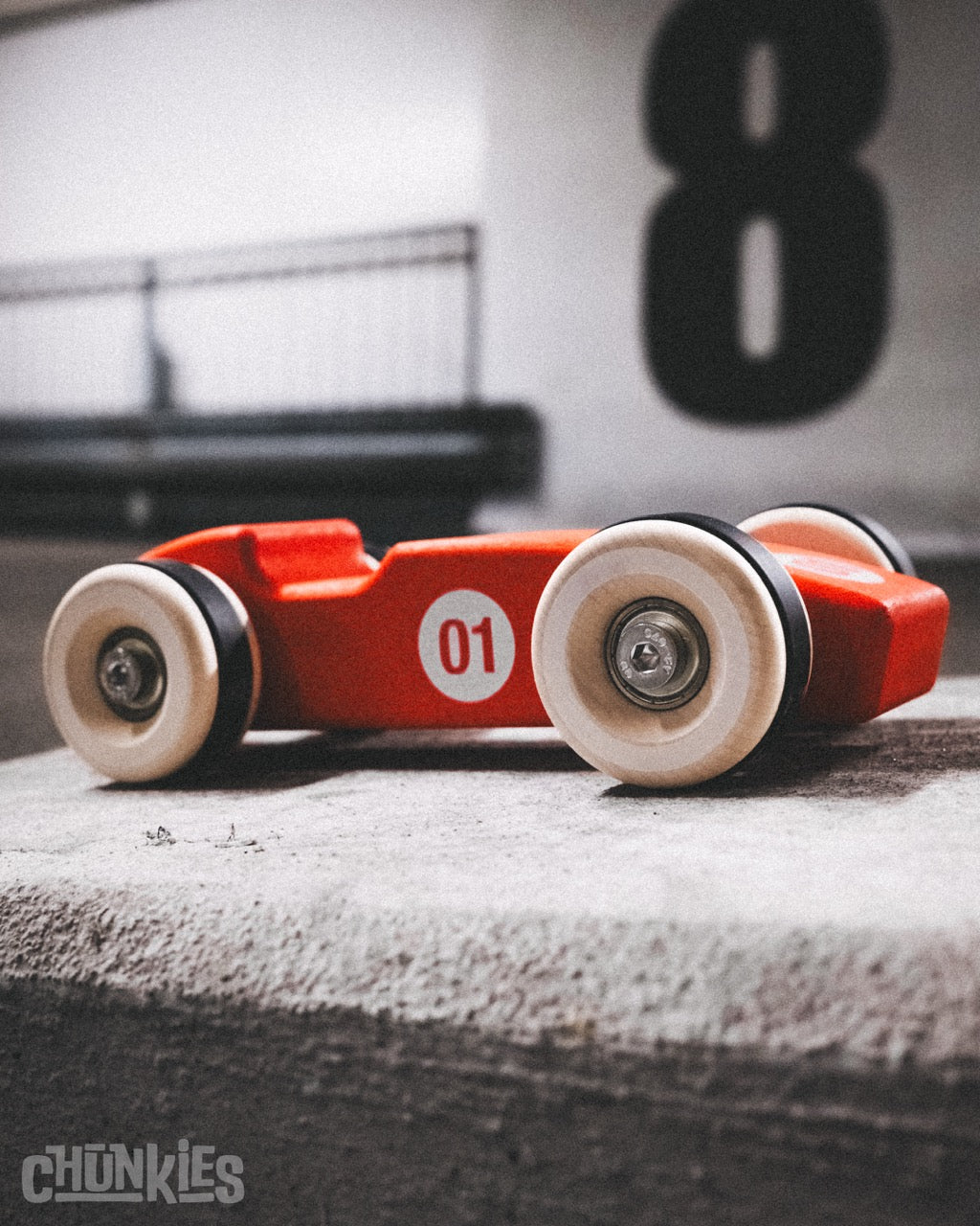 Red Chunkies toy race car with big wooden wheels and a number 1 on a concrete surface, blurred parking garage background. Ready to conquer your imagination and to pass on to the next generation. Heirloom quality