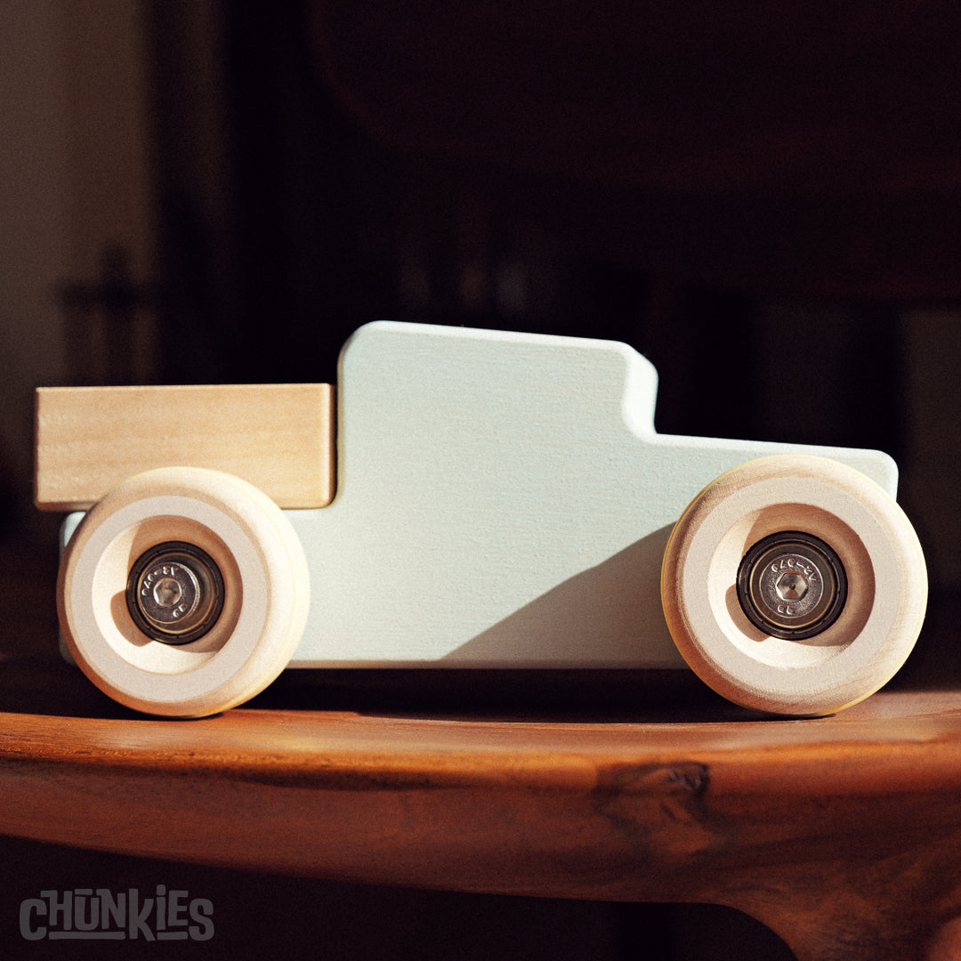 Side view of a Chunkies wooden toy truck with a light blue body and light wooden wheels on a wooden surface. A closer look at the stainless steel hardware and the high quality skate bearings for a smooth ride.
