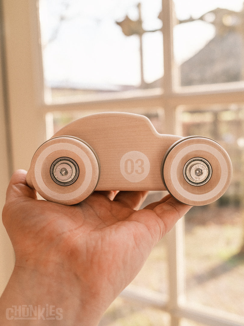 Chill Pepper Chunkies wooden toy car held in a hand with a blurred indoor background. With its natural maple wood, German quality and speedy skate bearings this vintage styled car is like no other.